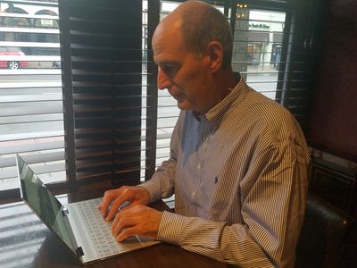A man sitting at a table typing on a laptop, involved in The Friday Baking Project.