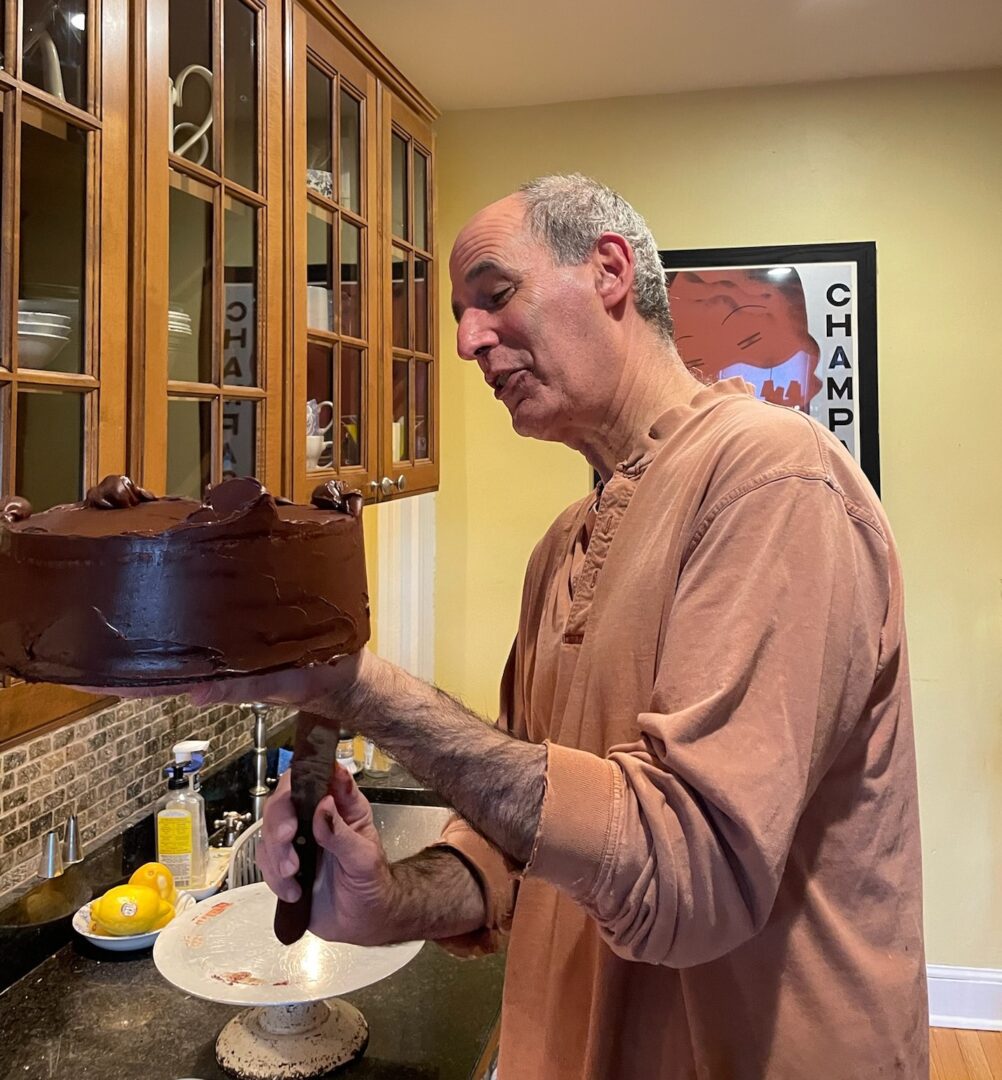 The Friday Baking Project features a man holding up a chocolate cake in a kitchen.