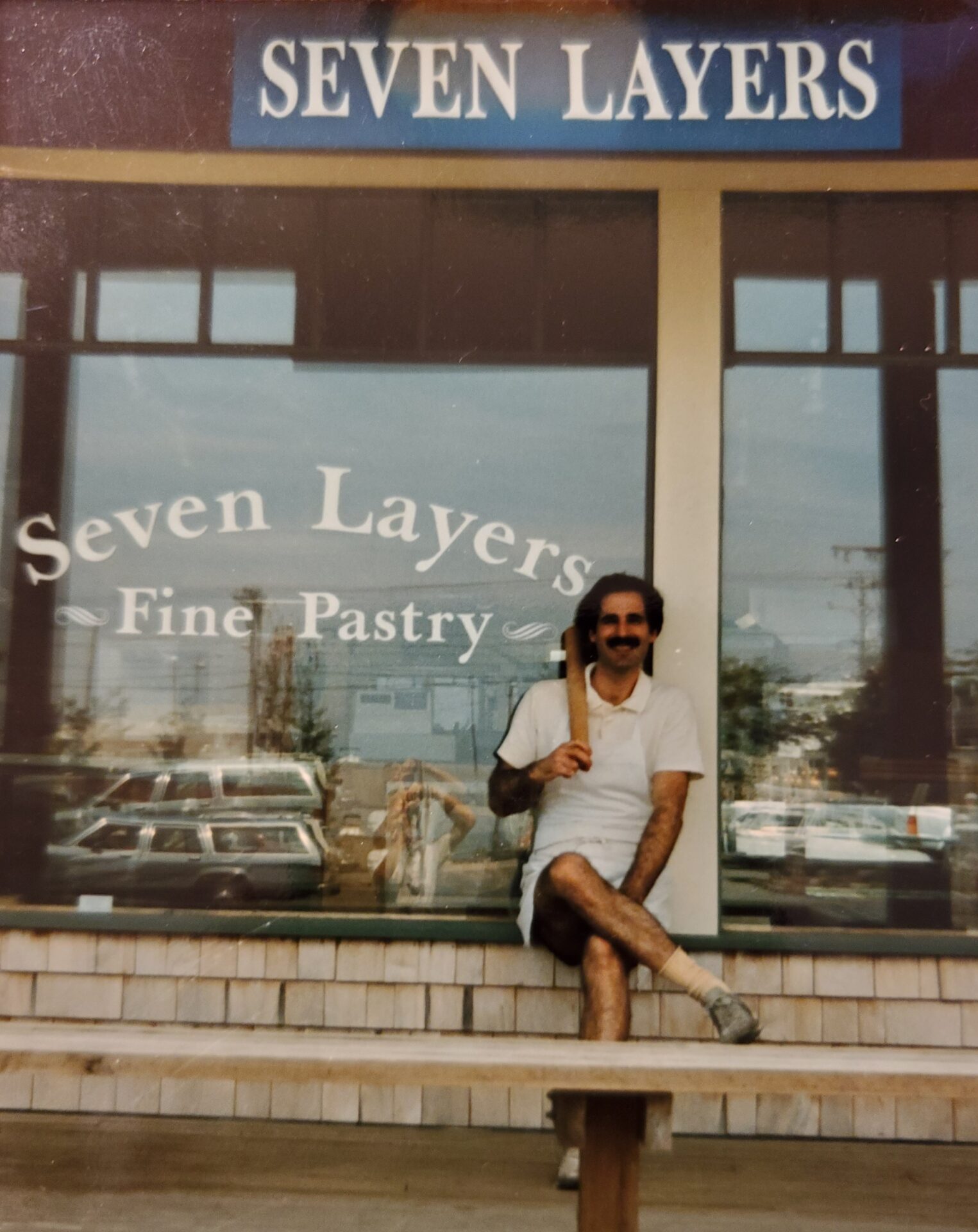 A man participating in The Friday Baking Project, sitting on a bench in front of a bakery.