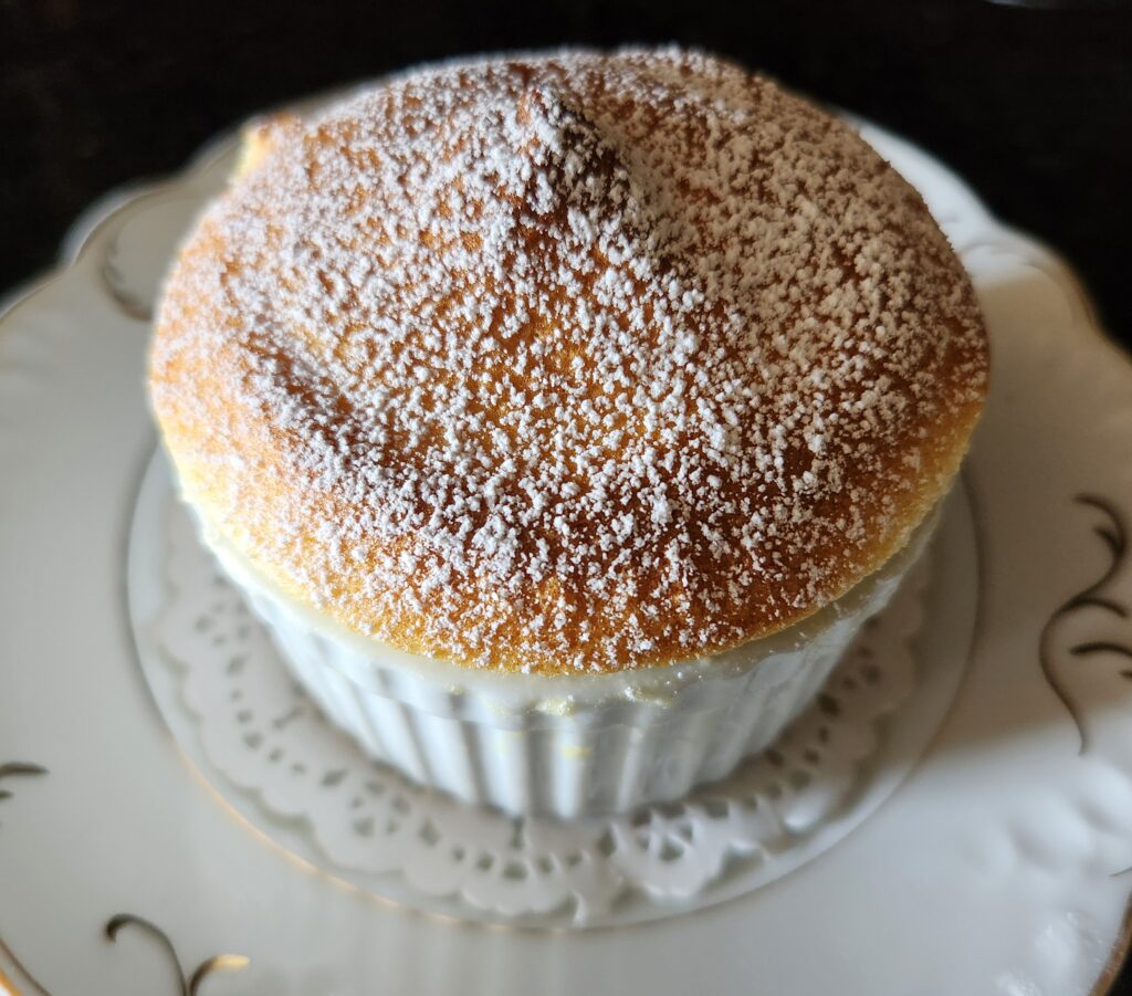 A freshly baked soufflé topped with powdered sugar, served in a white ramekin on an elegant patterned plate.