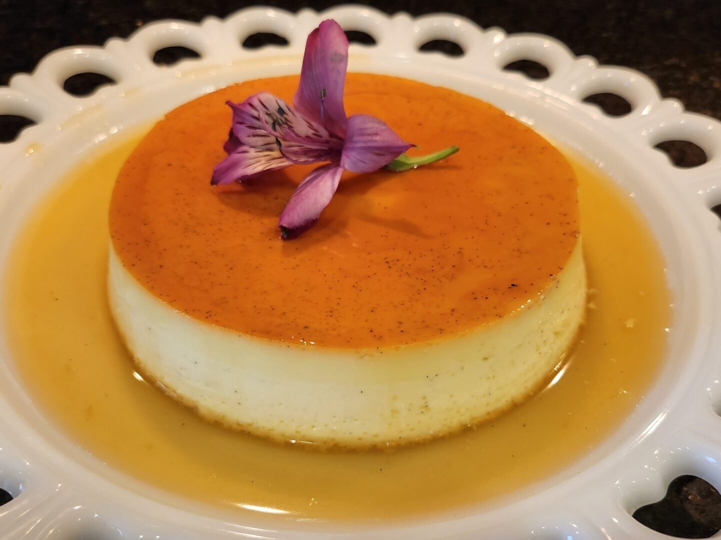 A flan topped with a purple flower on a white decorative plate, featured in The Friday Baking Project blog.