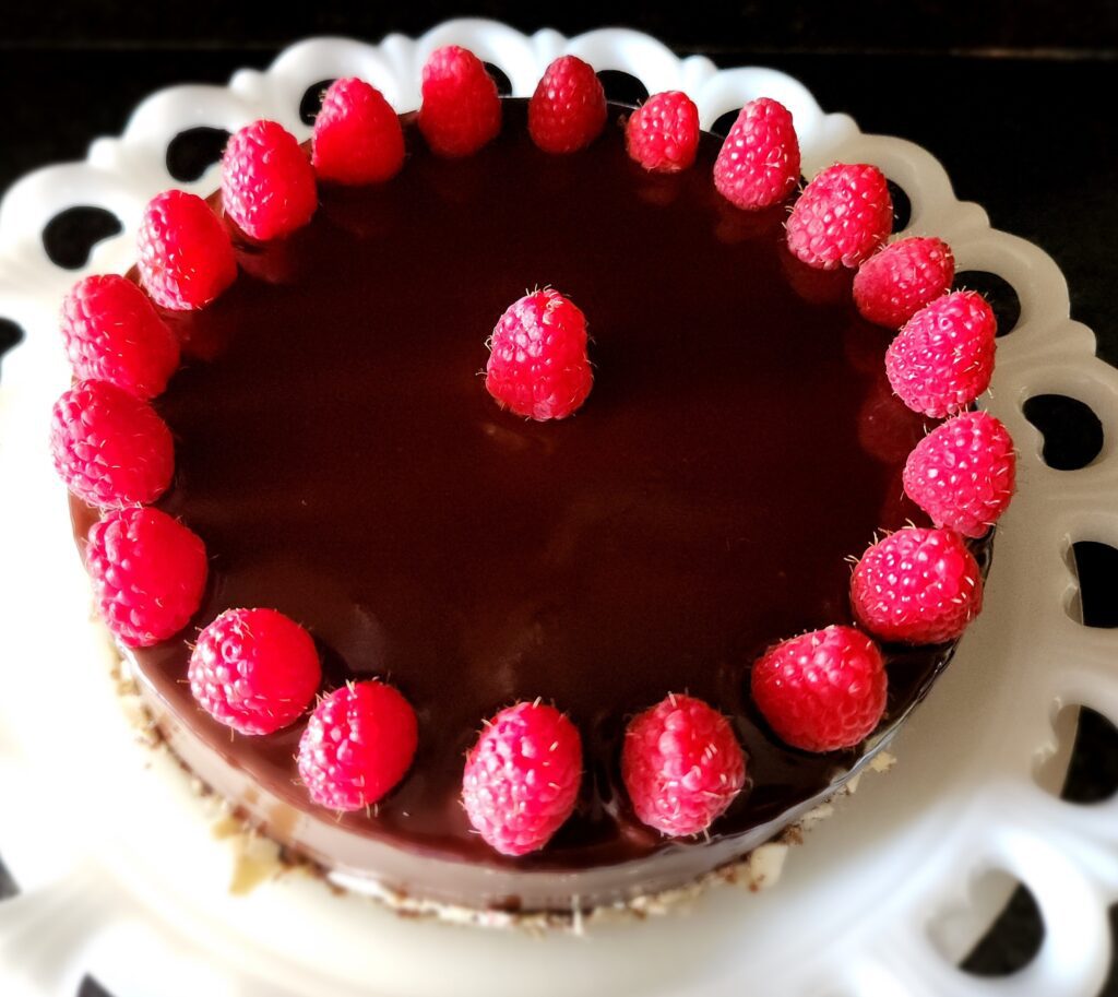 A chocolate ganache cake topped with fresh raspberries arranged in a circular pattern on a white decorative plate.